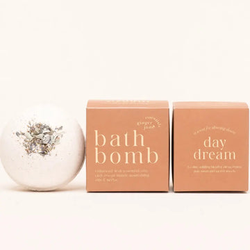 A white bath bomb by Ginger June in Daydream with herbs on top is shown next to two peach-colored boxes, one labeled 'bath bomb' and the other 'day dream', on a clean white background.
