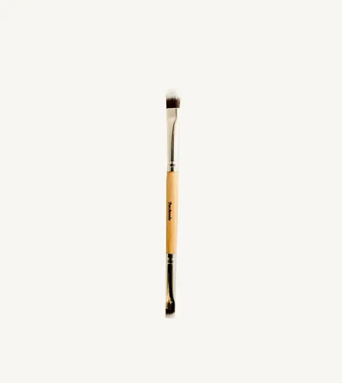 A photo of Pure Anadas vegan Duo Makeup Brush with one side that is a soft, feathery brush and the other an angled brush with a wooden, natural handle on a tan background.