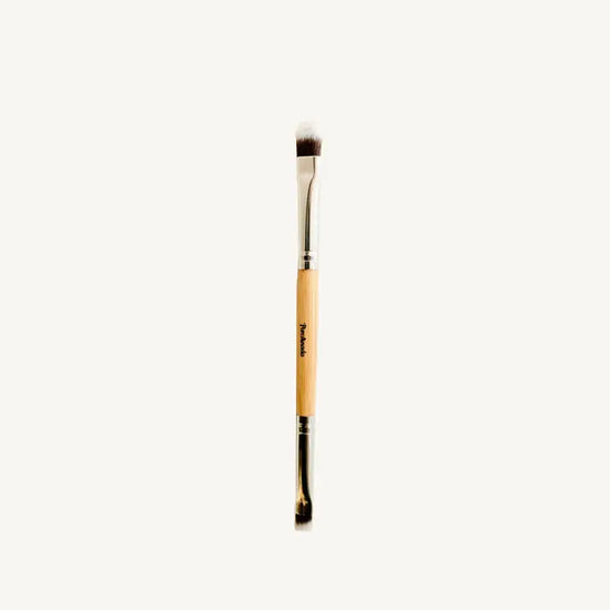 A photo of Pure Anadas vegan Duo Makeup Brush with one side that is a soft, feathery brush and the other an angled brush with a wooden, natural handle on a tan background.