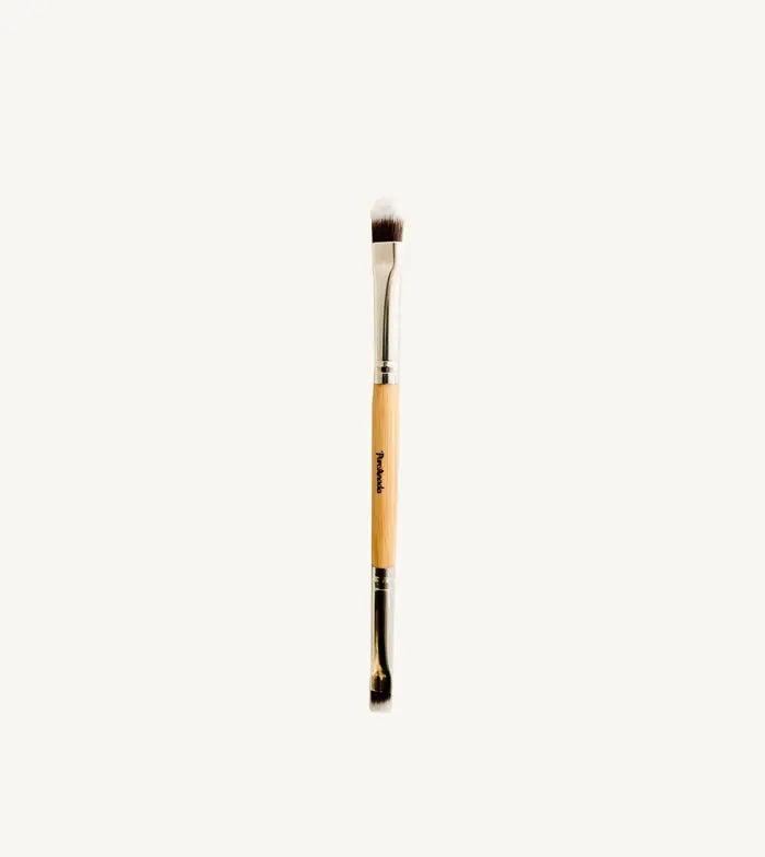 A photo of Pure Anadas vegan Duo Makeup Brush with one side that is a soft, feathery brush and the other an angled brush with a wooden, natural handle on a tan background.