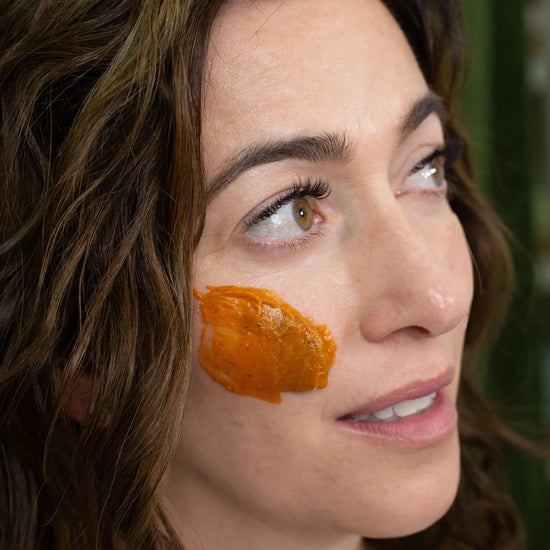 A woman with Simple Body Pumpkin Enzyme Mask on her cheek which is a bright orange texture on a green background.