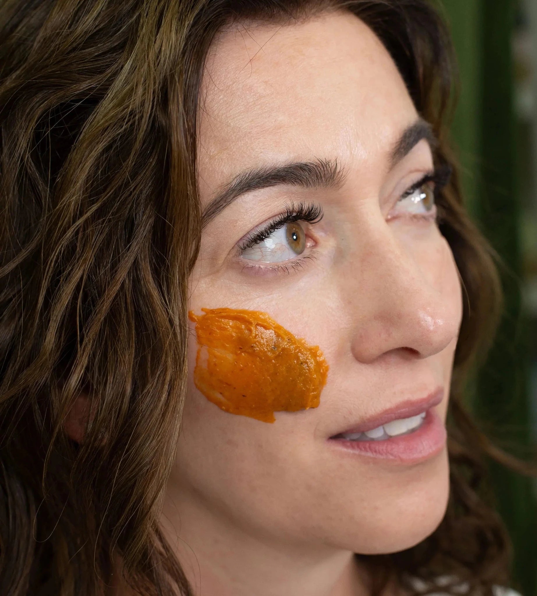 A woman with Simple Body Pumpkin Enzyme Mask on her cheek which is a bright orange texture on a green background.