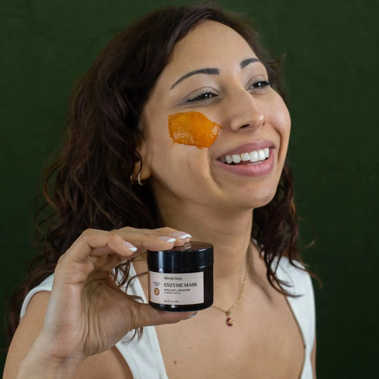 A woman looking off to the side with a swipe of Simple Body Pumpkin Enzyme Mask on her cheek which is a bright orange color holding a jar of the enzyme mask in one hand smiling with a dark green background.