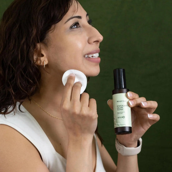 A woman looking sideways holding a bottle of Simple Body Exfoliate and Glow Tonic and holding a cotton pad in the other, smiling against a green background.