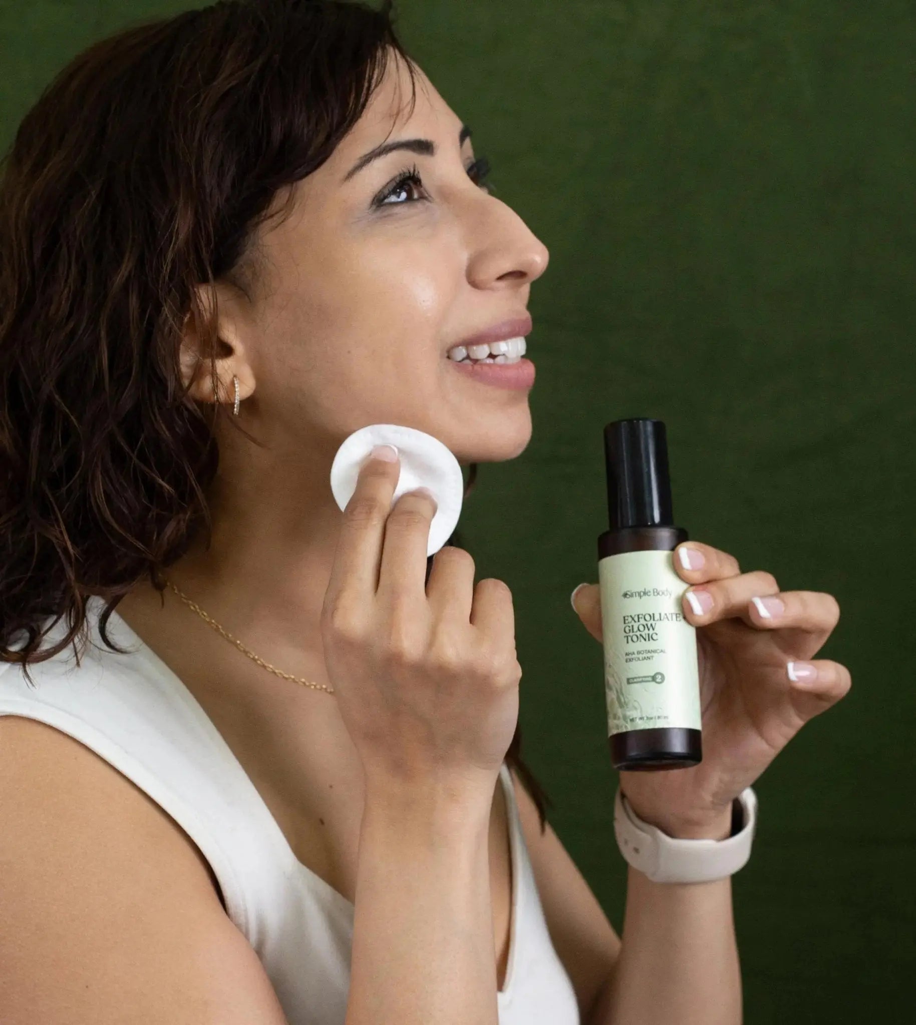 A woman looking sideways holding a bottle of Simple Body Exfoliate and Glow Tonic and holding a cotton pad in the other, smiling against a green background.