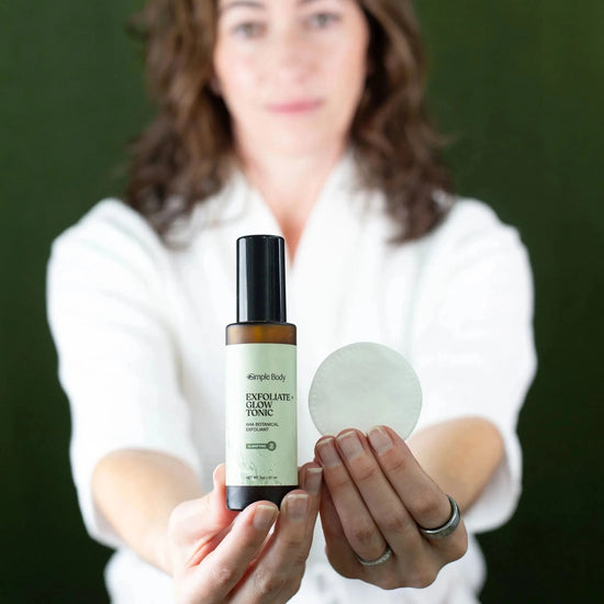 Woman holding a bottle of Simple Body Exfoliate and Glow with a cotton pad in the other hand against a dark green background