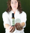 Woman holding a bottle of Simple Body Exfoliate and Glow with a cotton pad in the other hand against a dark green background