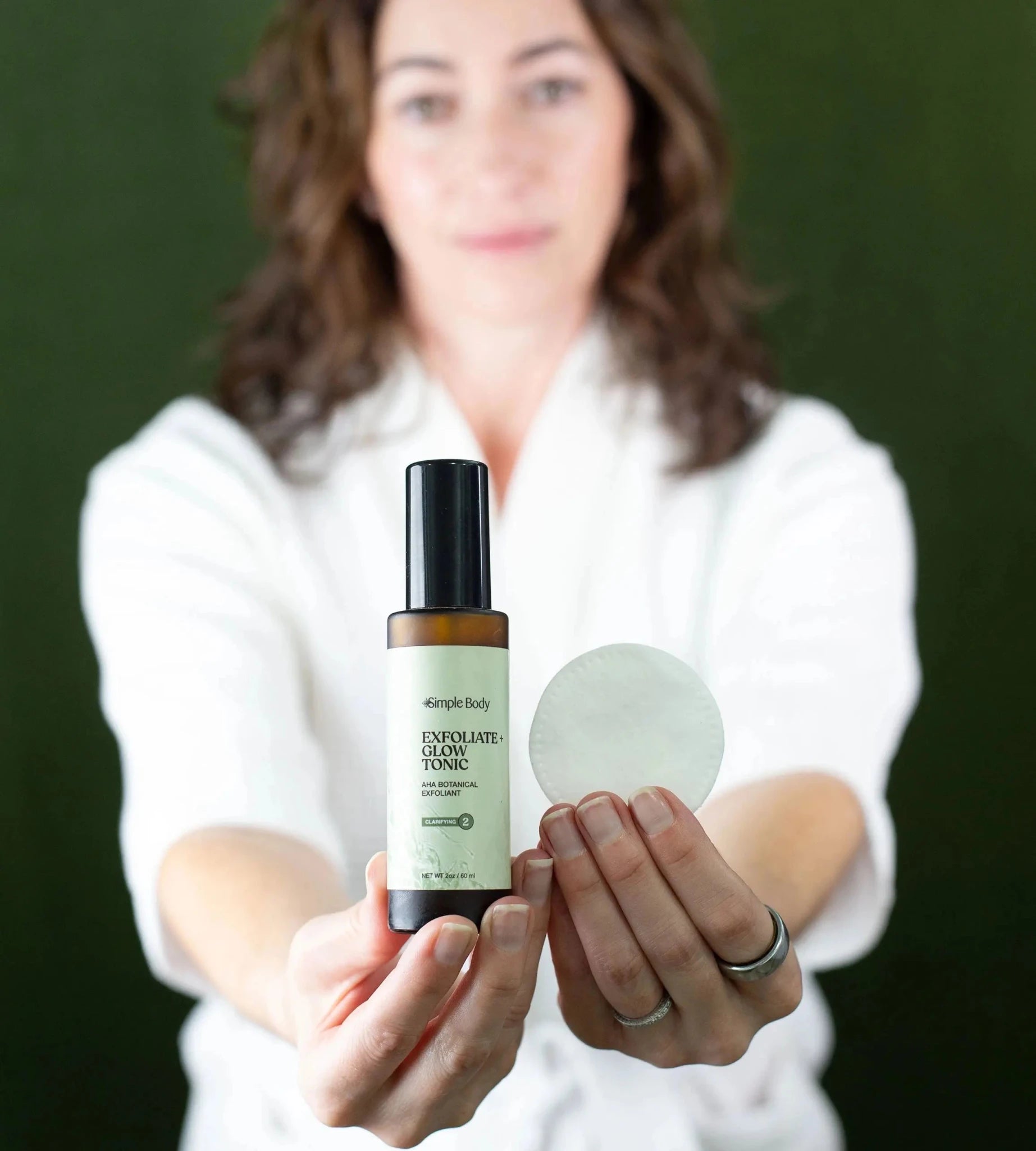 Woman holding a bottle of Simple Body Exfoliate and Glow with a cotton pad in the other hand against a dark green background