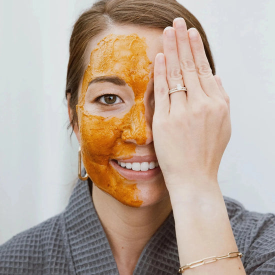 Shannon demonstrates a Simple Body Pumpkin Enzyme Mask on half her face which is an orange color holding her hand over the other half of her face in a gray robe on a white background.