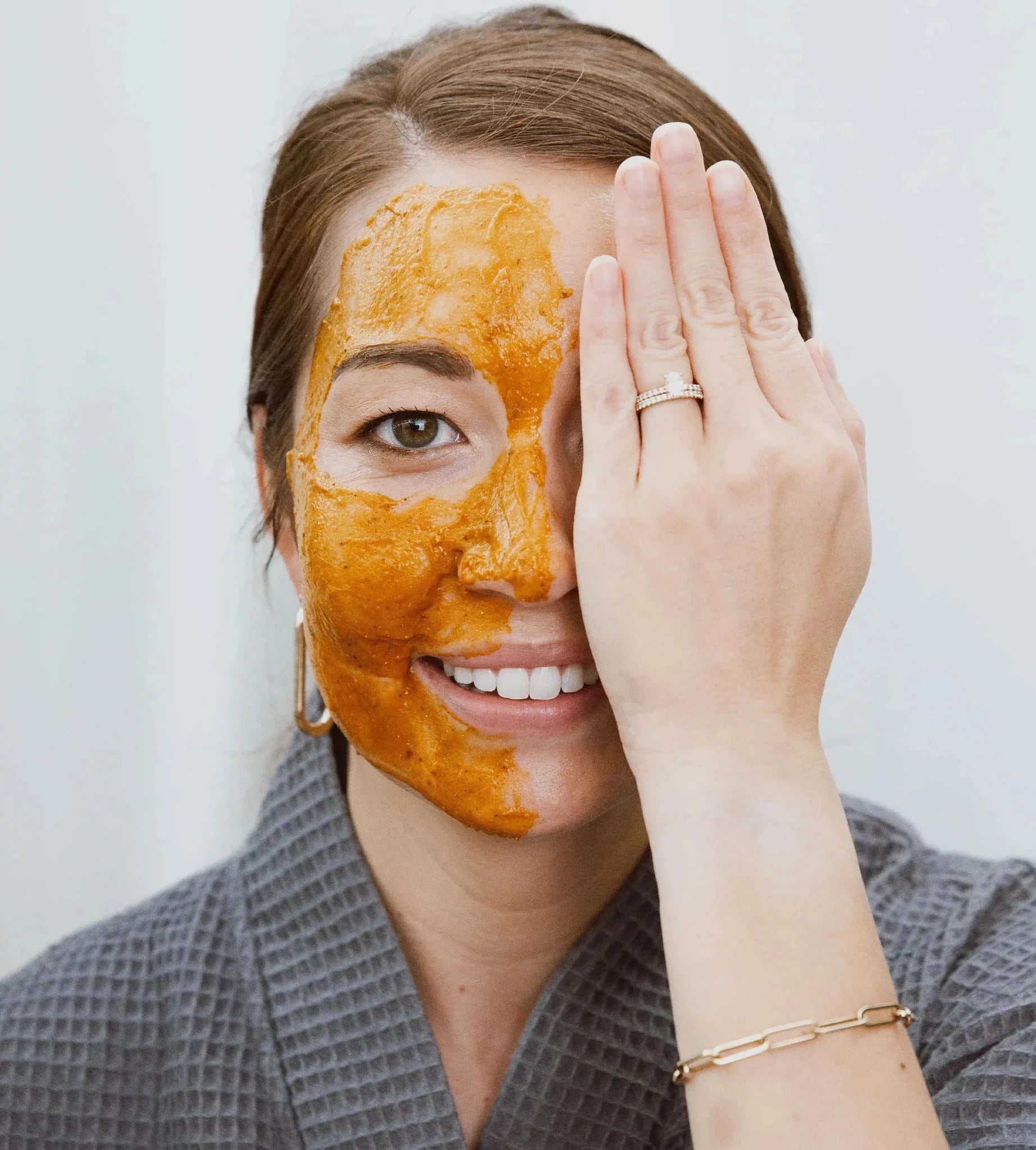 Shannon demonstrates a Simple Body Pumpkin Enzyme Mask on half her face which is an orange color holding her hand over the other half of her face in a gray robe on a white background.