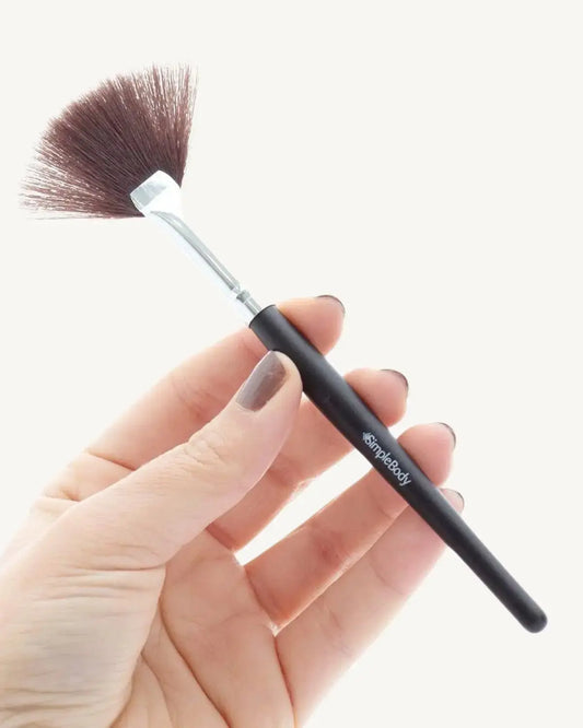 A hand holding a sleek black fan brush with synthetic bristles and a Simple Body logo printed on the handle.