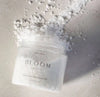A clear glass jar of Bloom Bath Co. bath soak in Wild Rose and Sandalwood with white bath salt, placed on a light gray textured surface with scattered white salt around it.