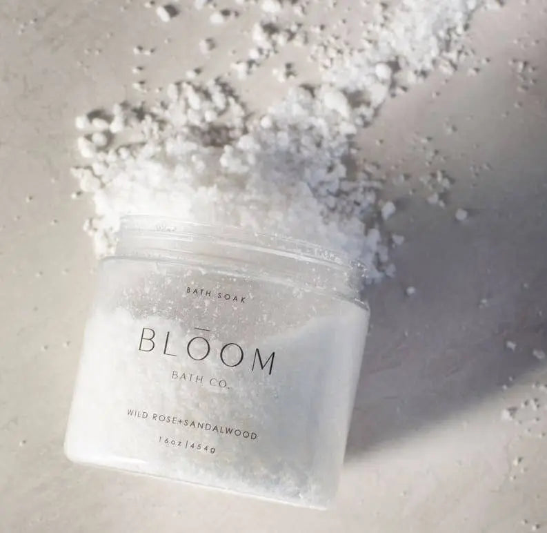 A clear glass jar of Bloom Bath Co. bath soak in Wild Rose and Sandalwood with white bath salt, placed on a light gray textured surface with scattered white salt around it.