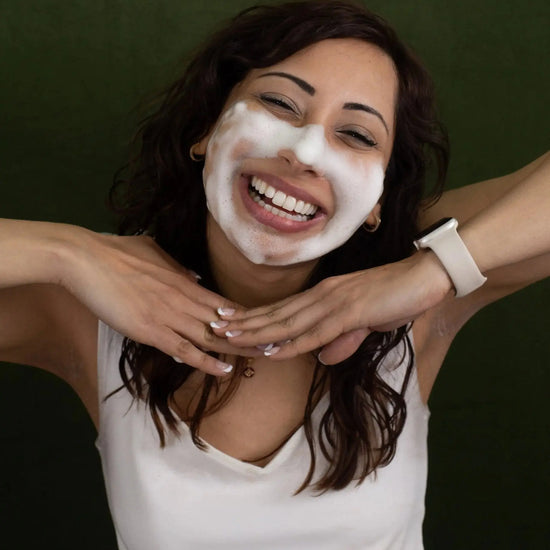 A woman with thick Simple Body Foaming Face Wash applied looking at the camera and smiling with a dark green background.