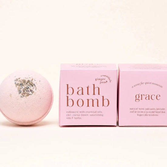 A pink bath bomb by Ginger June Bath Co with dried herbs on top center sits next to two pink boxes labeled 'bath bomb' and 'grace' on a clean white background.