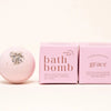 A pink bath bomb by Ginger June Bath Co with dried herbs on top center sits next to two pink boxes labeled 'bath bomb' and 'grace' on a clean white background.