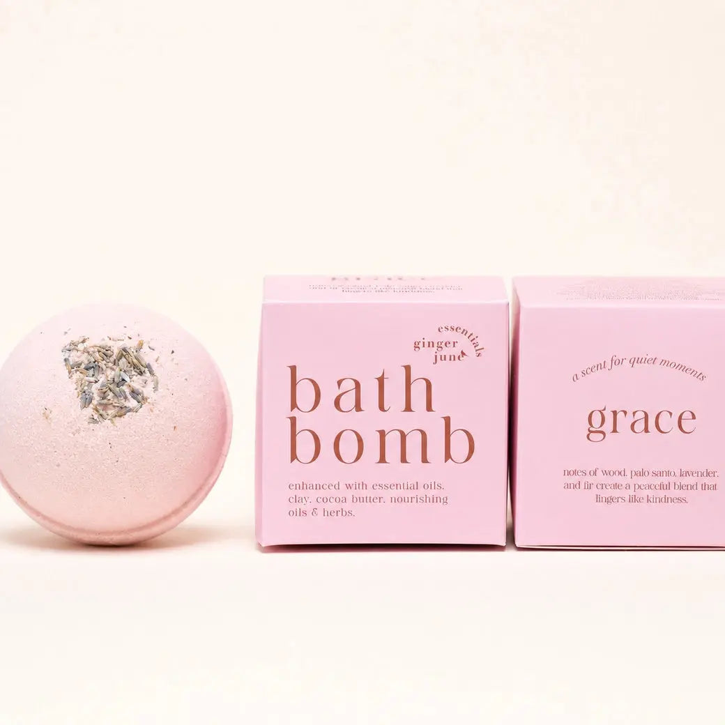 A pink bath bomb by Ginger June Bath Co with dried herbs on top center sits next to two pink boxes labeled 'bath bomb' and 'grace' on a clean white background.