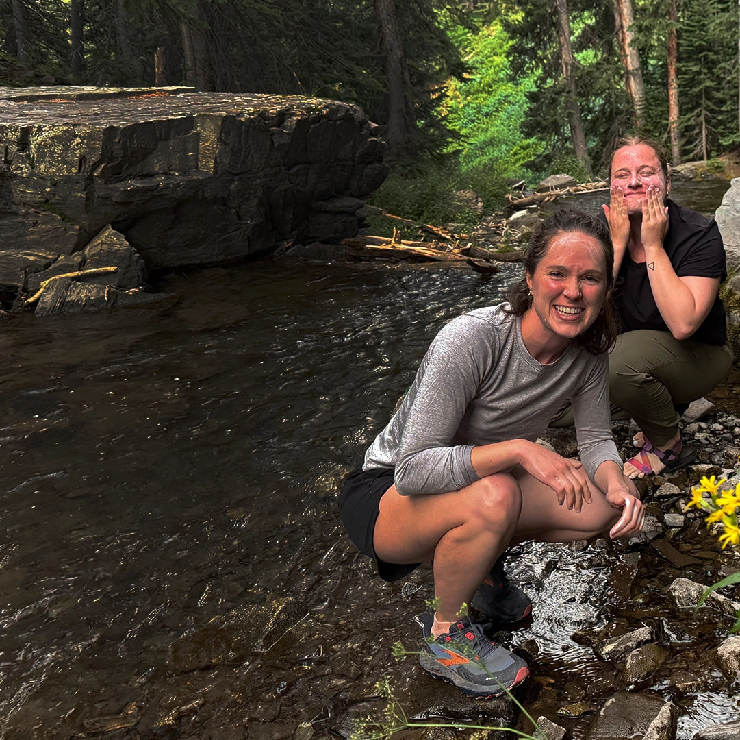 Two young ladies wash their face in a mountain creek with Simple Body Foaming Face wash while surrounded by nature.