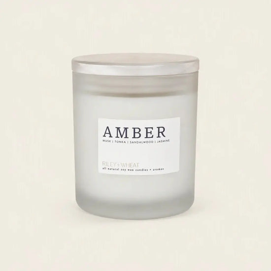 A frosted glass jar candle with a silver lid and a white label reading 'AMBER' and 'RILEY+WHEAT', photographed on a plain warm off-white background.