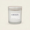 A frosted glass jar candle with a silver lid and a white label reading 'AMBER' and 'RILEY+WHEAT', photographed on a plain warm off-white background.