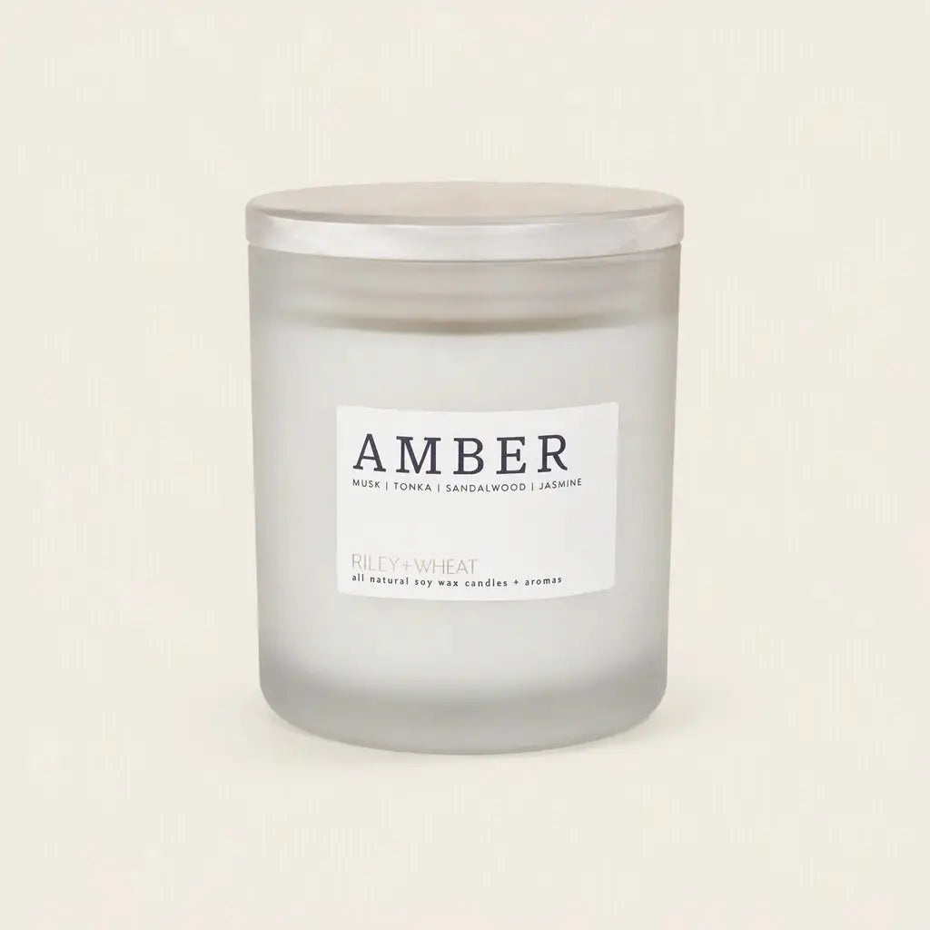 A frosted glass jar candle with a silver lid and a white label reading 'AMBER' and 'RILEY+WHEAT', photographed on a plain warm off-white background.