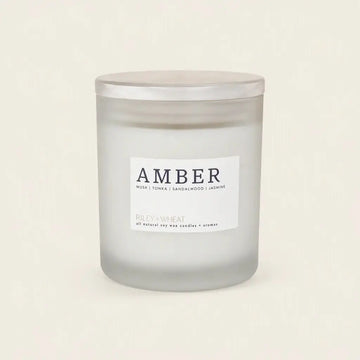 A frosted glass jar candle with a silver lid and a white label reading 'AMBER' and 'RILEY+WHEAT', photographed on a plain warm off-white background.