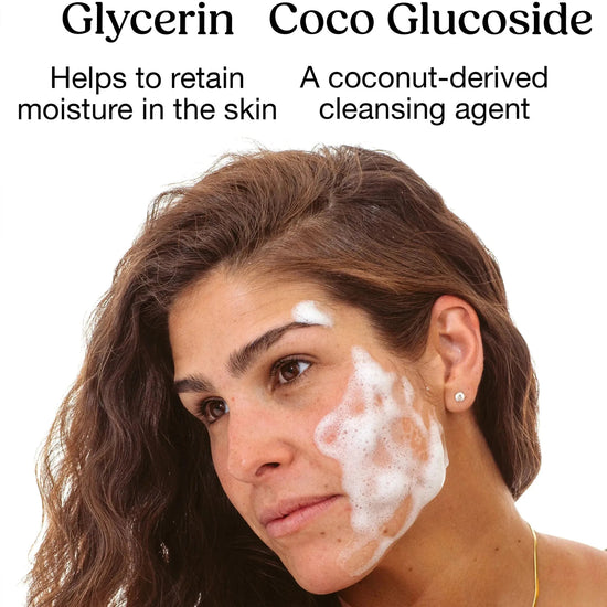 A young woman looks to the side with long dark hair and Simple Body Foaming Face Wash applied to her cheek on a white background with Glycerin and Coco Glucoside called out as key ingredients.