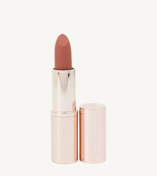 A photo of Pure Anada's Carefree Matte Lipstick in a rose gold tube and a warm orange - rust color on a tan background.