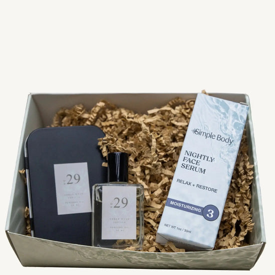 A cute gift box that features a black tin with the No. 29 on it and a perfume bottle next to a Simple Body Nightly Face Serum on kraft paper in a gift box.