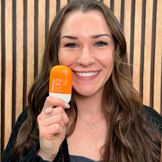 Woman holding a bottle of Think Sun Serum sunscreen with a wooden panel background