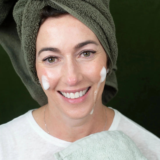 A woman with a green towel on her head and Simple Body Foaming Face Wash on her cheeks against a green background.