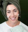 A woman with a green towel on her head and Simple Body Foaming Face Wash on her cheeks against a green background.