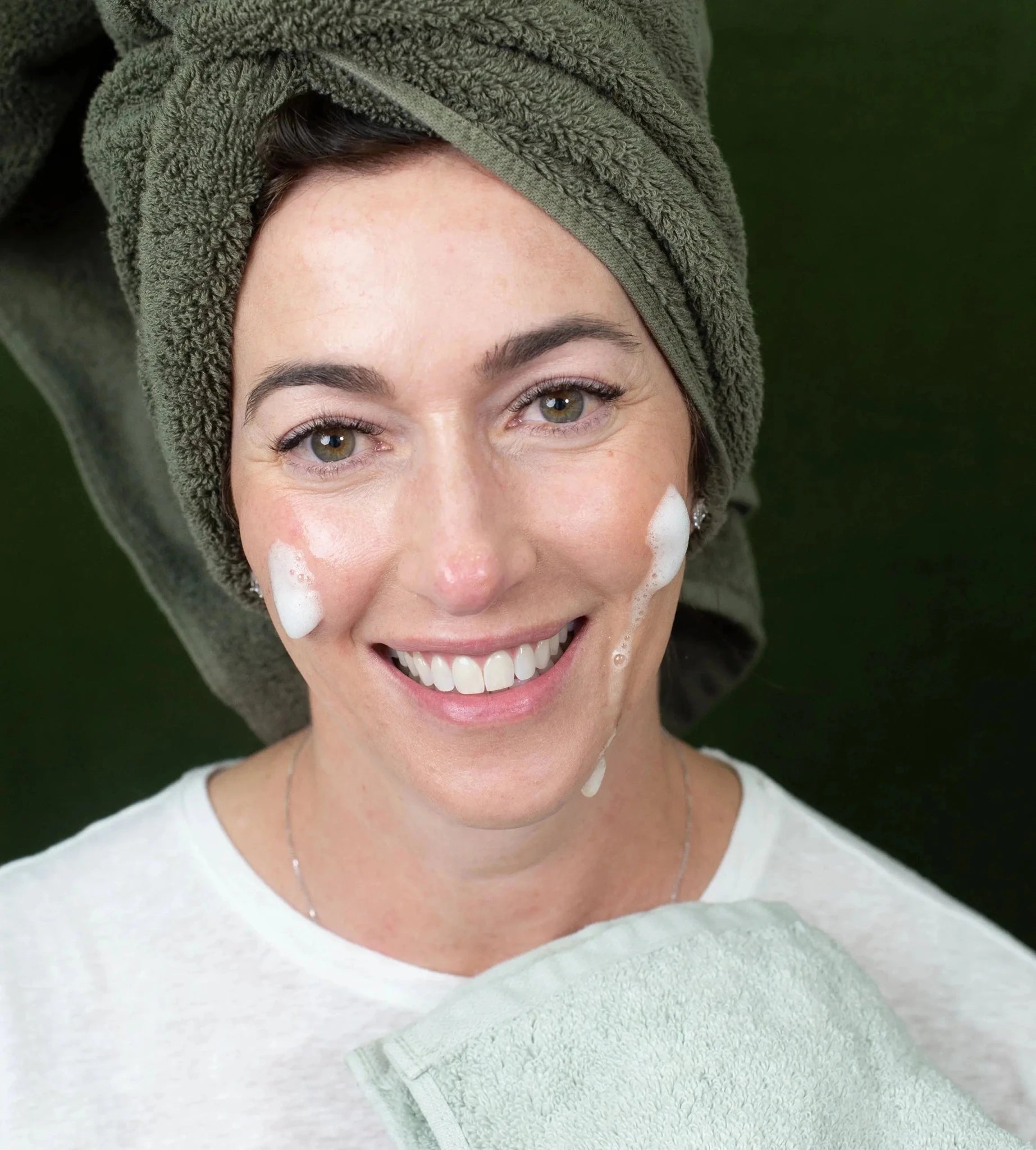 A woman with a green towel on her head and Simple Body Foaming Face Wash on her cheeks against a green background.