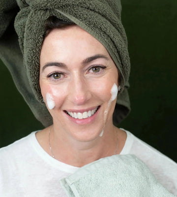A woman with a green towel on her head and Simple Body Foaming Face Wash on her cheeks against a green background.