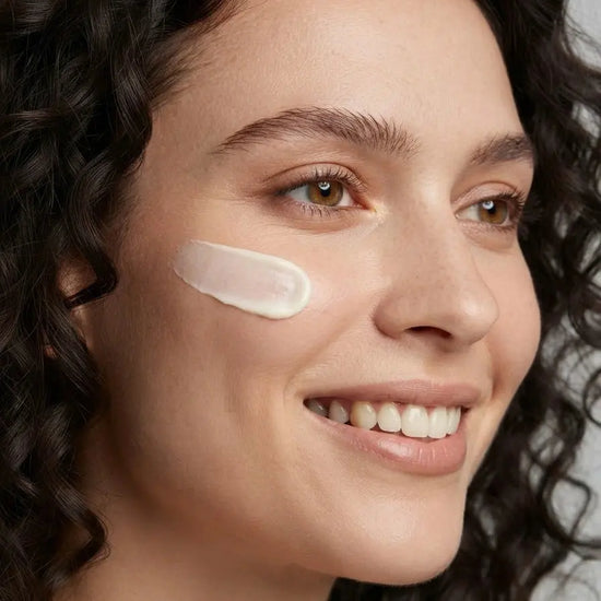 A woman with black curly hair smiles and has a swipe of Simple Body's Barrier Defense Serum on her cheek which is a white, creamy texture.