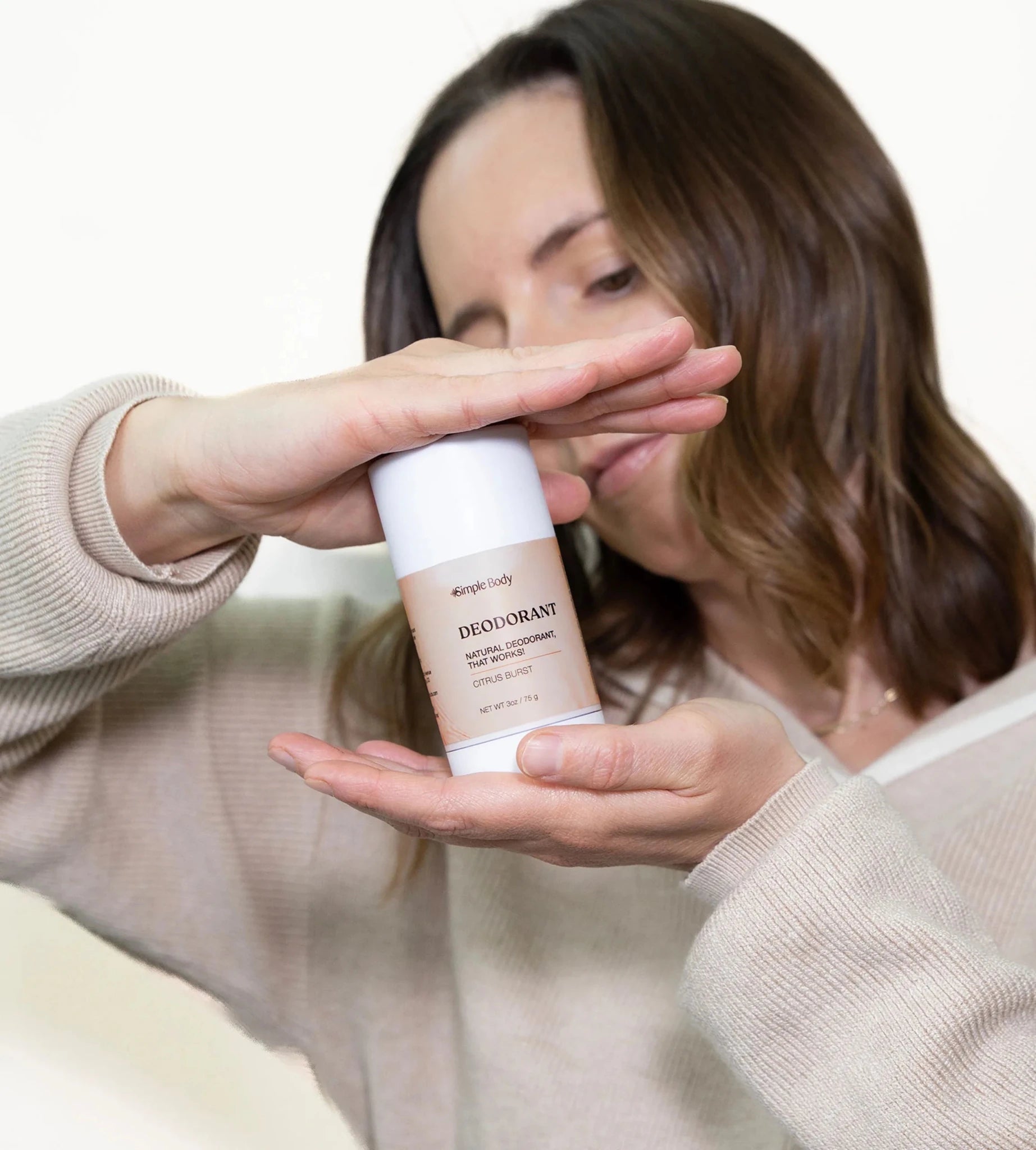 A beautiful young mom with brown hair holds a stick of Simple Body Natural Deodorant, Citrus Burst between her two hands.