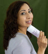 Woman holding a jar of Simple Body All Natural Lavender Deodorant looking over her shoulder with a purple sweater and a dark green background.
