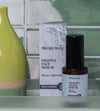 A bottle of Nightly Facial Serum from Simple Body sits on a bathroom countertop with a lime green vase and green tile background.