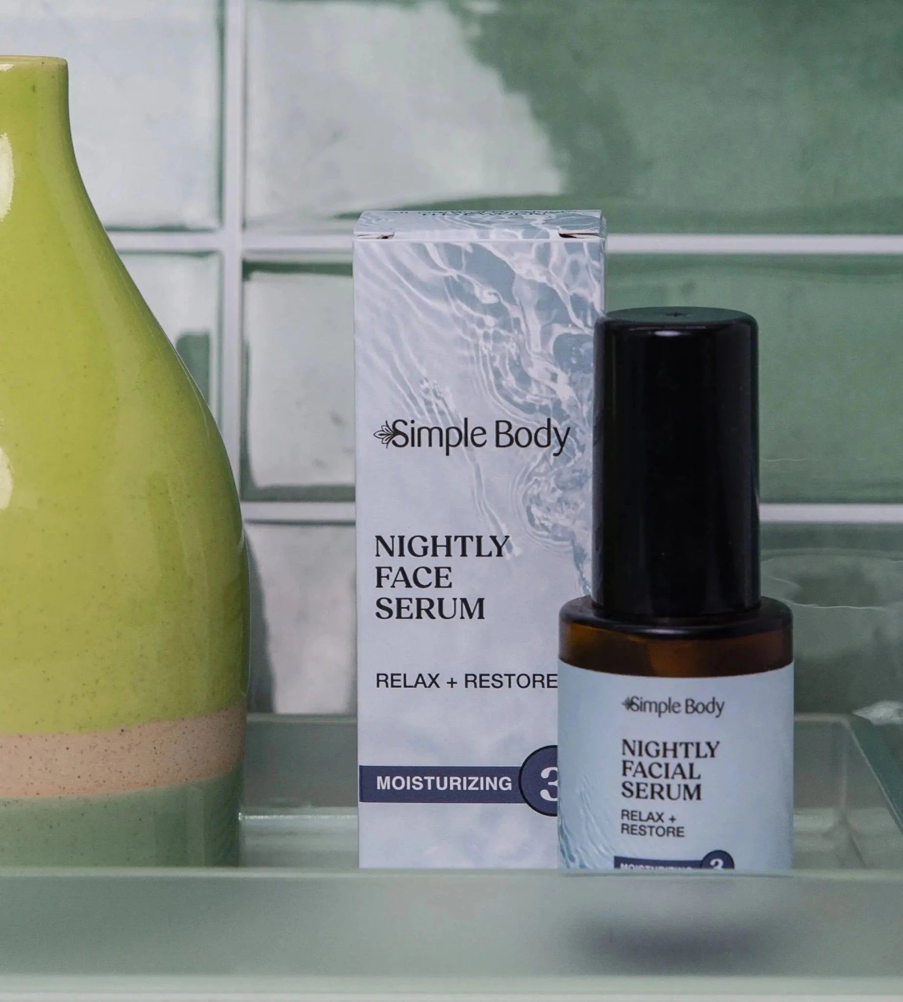 A bottle of Nightly Facial Serum from Simple Body sits on a bathroom countertop with a lime green vase and green tile background.