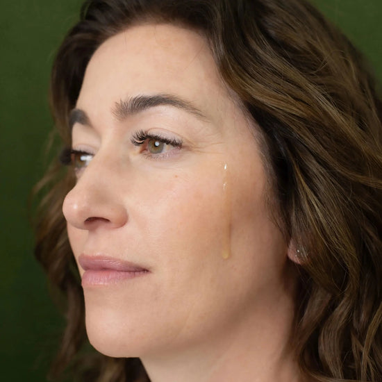 Close-up of a woman's face with a drop of Simple Body Pomegranate Oil dripping down her cheek with a green background