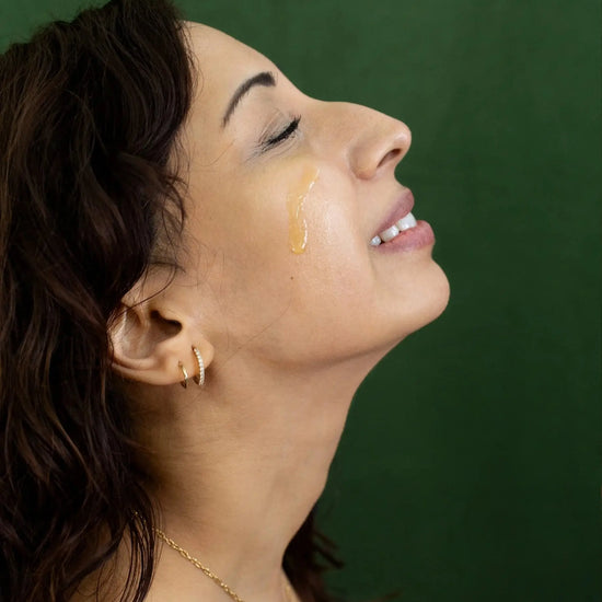 A woman looking up with her eyes closed and a drip of Simple Body Pomegranate Facial Serum dripping down her cheek with a dark green background.