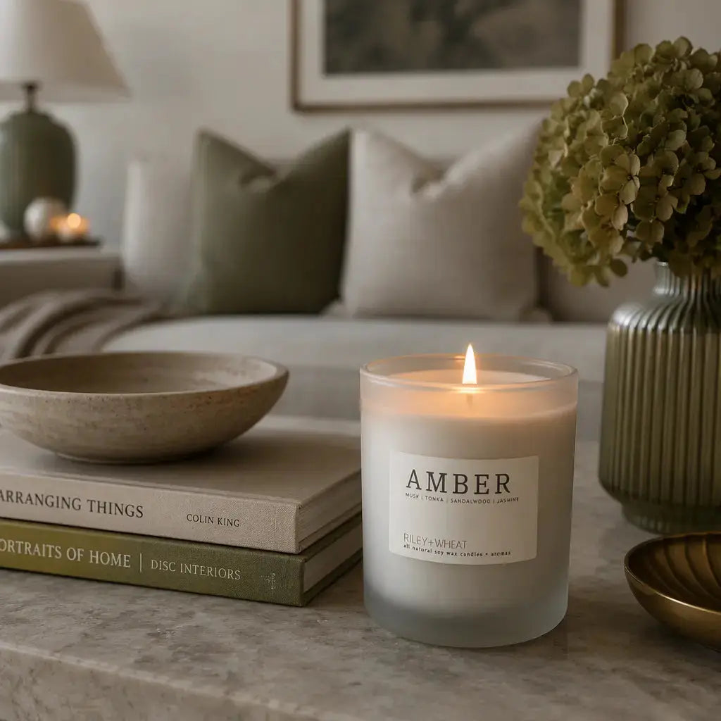 A lit scented candle in a frosted glass jar sits on a marble surface in a styled living room setting, surrounded by books, a ceramic bowl, a vase with green hydrangeas, and a sofa in the background. The candle label reads 'AMBER' with smaller text 'SILKY WHEAT' and 'ORLANDO, FL'.