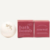 A white round bath bomb by Ginger June Bath Co with herbs on top sits beside two dusty rose product boxes labeled 'bath bomb' and 'renewal' on a warm cream background.