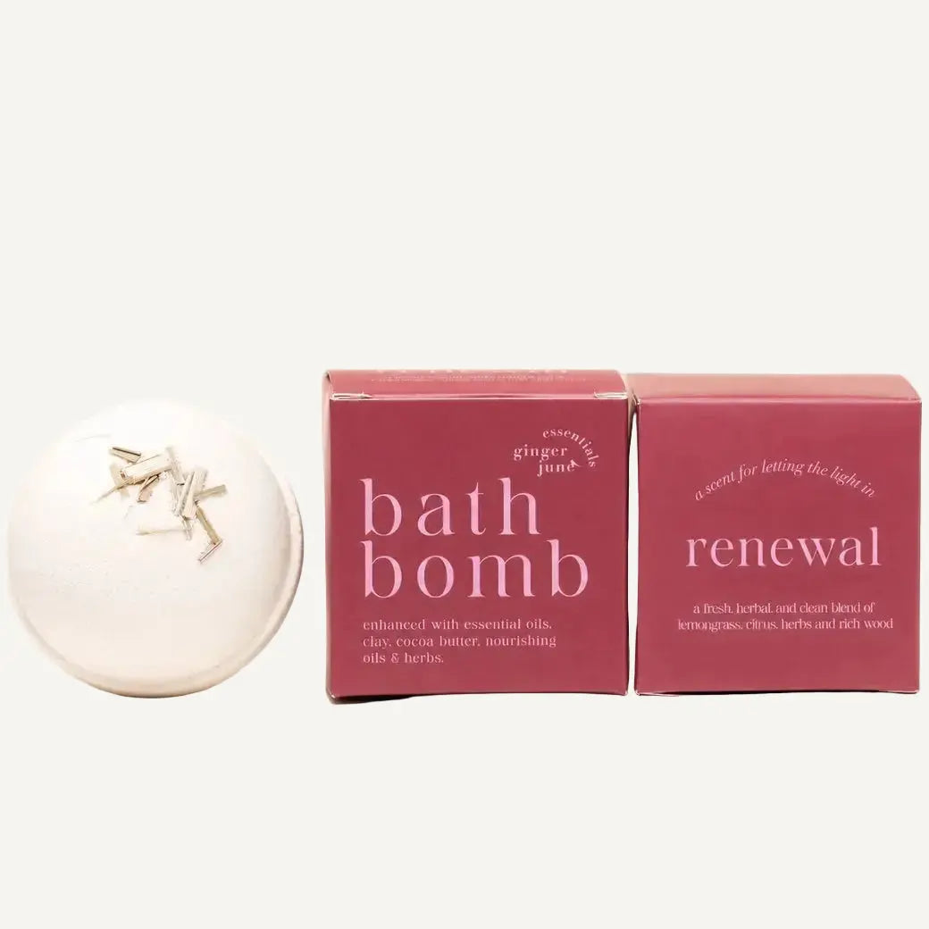 A white round bath bomb by Ginger June Bath Co with herbs on top sits beside two dusty rose product boxes labeled 'bath bomb' and 'renewal' on a warm cream background.
