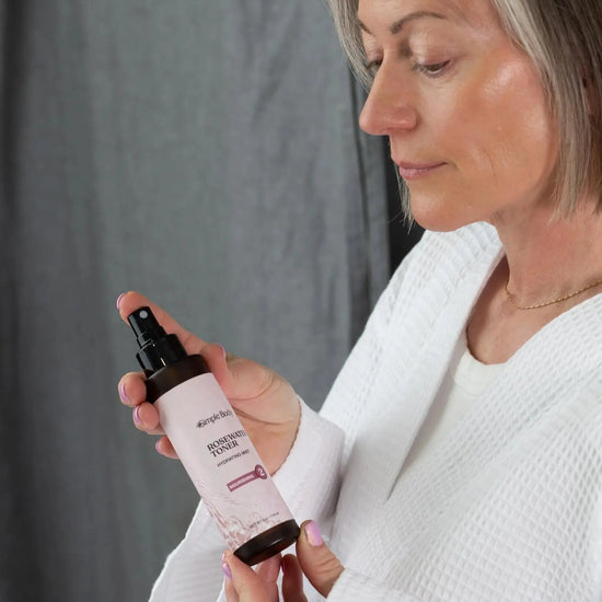 Woman holding a bottle of Simple Body Rosewater Facial Toner looking down at it in a white robe with a gray background.
