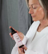 Woman holding a bottle of Simple Body Rosewater Facial Toner looking down at it in a white robe with a gray background.