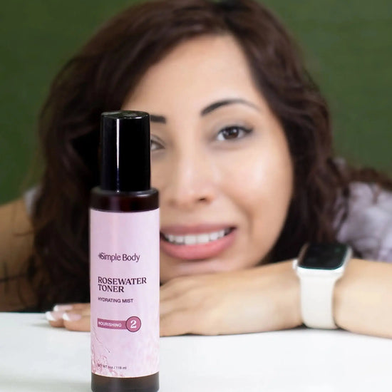 A woman looking directly at you but slightly blurred and a bottle of Simple Body Rosewater Facial Toner in front in focus with a green background.