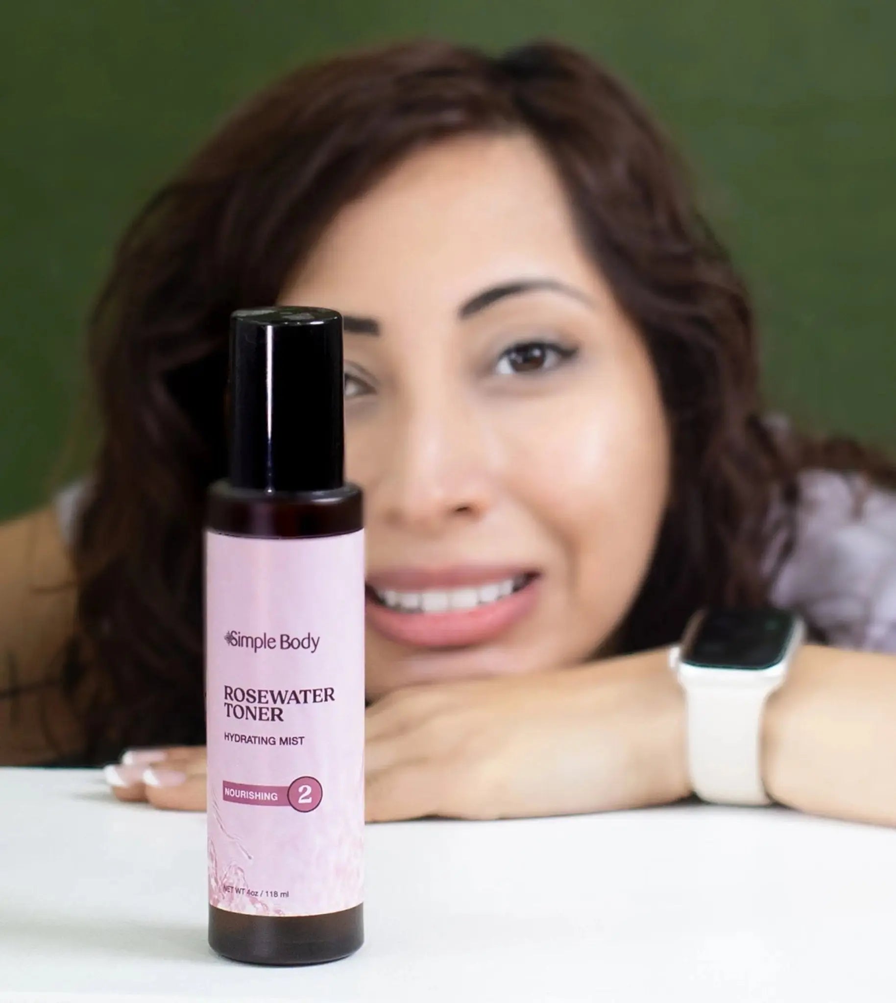 A woman looking directly at you but slightly blurred and a bottle of Simple Body Rosewater Facial Toner in front in focus with a green background.
