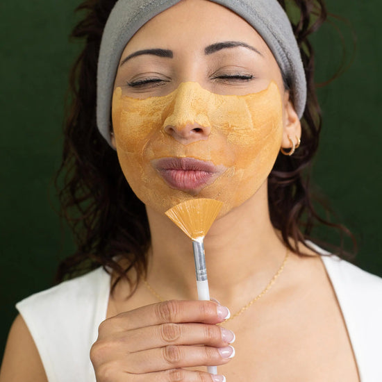 Woman applying a Simple Body Caramel Clay facial mask with a brush on a green background