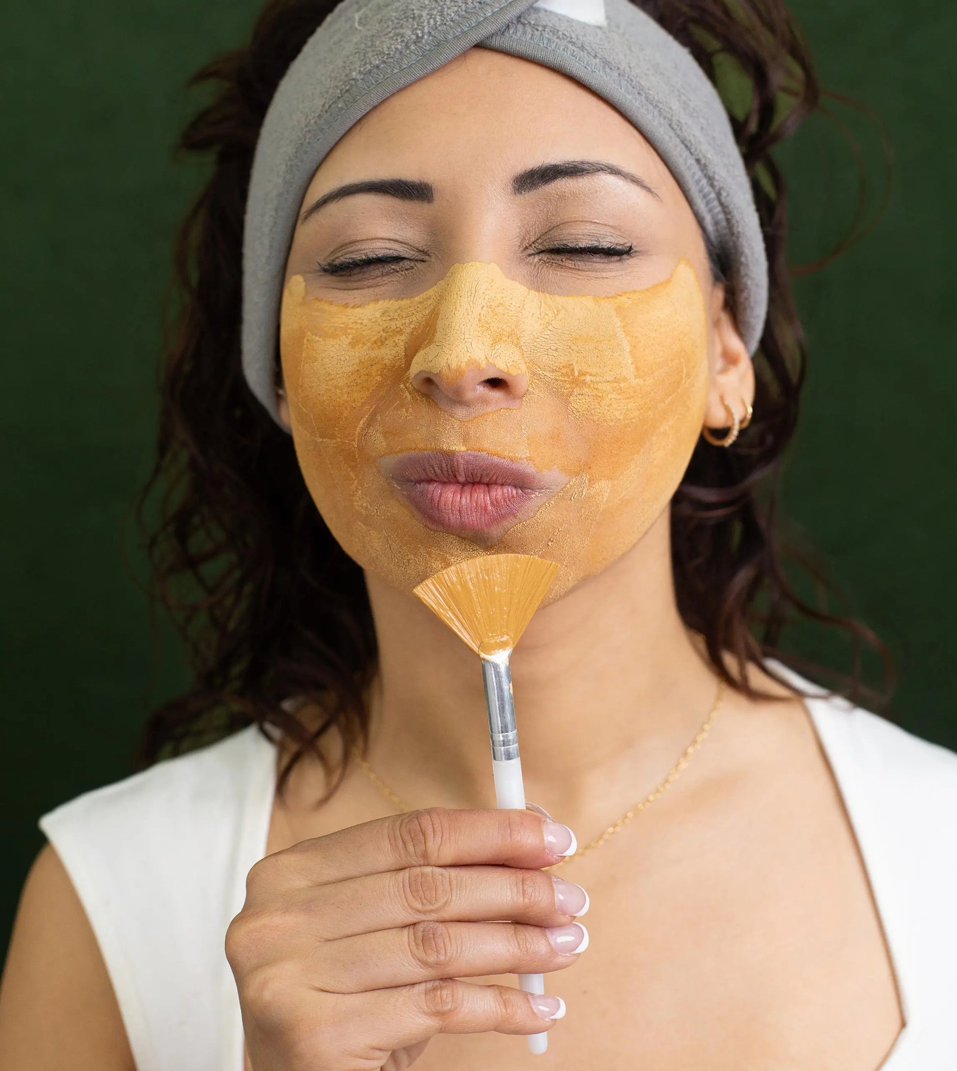 Woman applying a Simple Body Caramel Clay facial mask with a brush on a green background